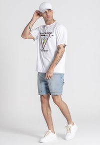 Biele grafické tričko s textom a dizajnom, modré denimové šortky a biele tenisky s béžovými šnúrkami. Doplnky zahŕňajú bielu šiltovku.