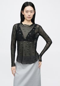 Čierny priesvitný top s dlhým rukávom s vystúpenou textúrou bodiek, hlbokým výstrihom do tvaru V a priliehavým strihom. Kombinovaný so svetlosivou sukňou.