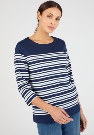 Femme aux cheveux attachés, portant un haut rayé bleu marine et blanc à manches longues et un jean bleu, debout devant un fond gris clair.