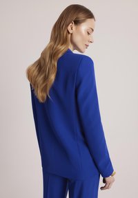 Dopasowana niebieska marynarka z eleganckimi liniami i gładką teksturą, zestawiona z pasującym garniturem, widok z tyłu. Widoczny akcent w postaci złotej bransoletki.