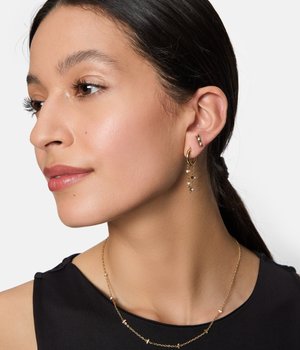 Orecchini a cerchio dorati con elementi pendenti e piccoli gemme trasparenti. Collana semplice a catena con forme circolari, indossata con un top nero.