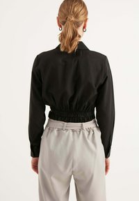 Giacca nera corta con maniche lunghe, vita elastica e colletto, abbinata a pantaloni grigio chiaro con vita arricciata.