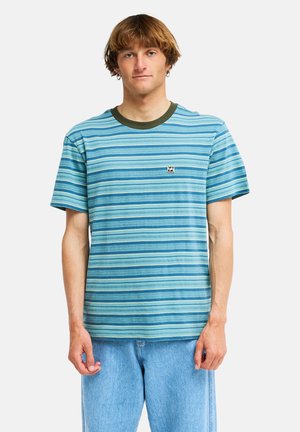 Jonge man die een blauw-groen gestreept T-shirt met korte mouwen en lichtblauwe spijkerbroek draagt, staand tegen een effen witte achtergrond.