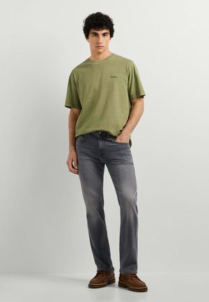 Giovane uomo che indossa una t-shirt verde oliva, jeans grigi e scarpe marroni, in piedi con una mano in tasca contro una parete chiara e semplice.