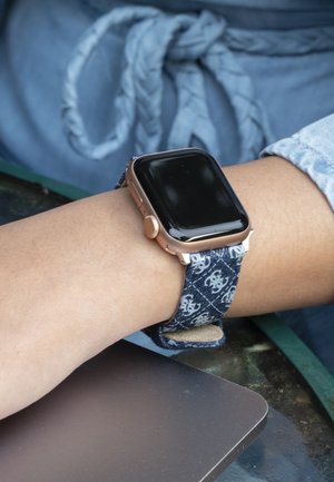 Pametna ura s črnim zaslonom na dotik, ohišjem rožnatega zlata in modrim tkanim paščkom z belim vzorcem. Roka počiva na površini.