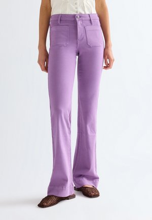 Vrouw draagt hoge taille, lavendelkleurige uitlopende broek met voorzakken en bruine gevlochten platte schoenen op een witte achtergrond.