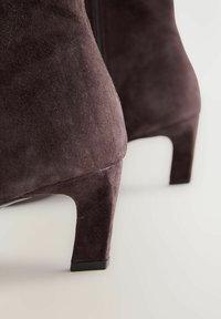 Botas tobilleras de ante morado oscuro con tacón cónico y cierre de cremallera, con una superficie texturizada y bordes suaves. Diseño minimalista.