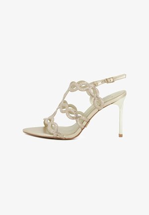 Sandalo beige con tacco alto con cinturini decorativi ad anello ornati da piccoli strass e fibbia regolabile alla caviglia.