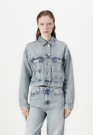 Giacca di jeans azzurra cropped con due tasche sul petto, chiusura con bottoni e una texture leggermente sbiadita. Abbinata a jeans blu coordinati.