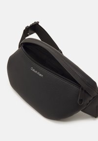 Musta nahkainen vyölaukku, jossa on teksturoitu pinta, vetoketjullinen suljin ja muovinen säädettävä hihna. Edessä hopeinen "Calvin Klein" -logo.