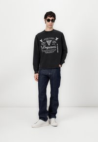 Sort sweatshirt med hvid grafisk tryk, blå jeans og hvide sneakers. Sweatshirten har rund hals og ribkantede ærmer. Solbriller fuldender looket.