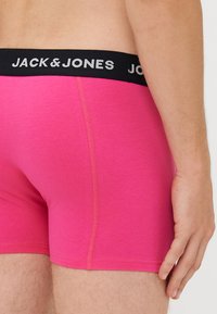 Roza bombažna bokserica z črnim elastičnim pasom, ki nosi napis "JACK & JONES" v belem tisku. Gladka tekstura in prilegajoča se zasnova.