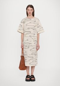 Beige bomulls kjole med abstrakte svarte linjer og grønne detaljer. Den har korte ermer, løs passform og en lomme foran. Bæres med en vevd brun veske.