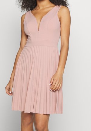 Kobieta w bezrękawowej sukience w kolorze przygaszonego różu, z głębokim dekoltem w szpic i plisowaną spódnicą, stojąca na jednolitym jasnym tle.