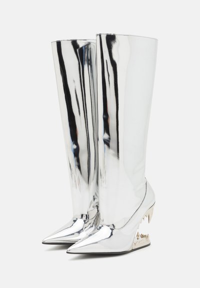 Silver knee-high boots with a reflective surface, pointed toe, and jagged metallic heel. Smooth texture with a sleek design.