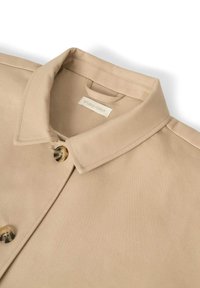 Beige Hemdkragen mit Knopfverschluss, aus glattem Stoff. Das Label "STUDIO FEDER" ist in der Nähe des Ausschnitts sichtbar.