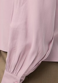 Manche longue bouffante en tissu rose clair avec poignet froncé et bouton, portée sur un pantalon beige.