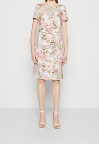 Kobieta w sukience w kwiaty o długości do kolan, z krótkim rękawem, w delikatnych odcieniach różu i beżu, zestawiona z białymi szpilkami na kostkę.