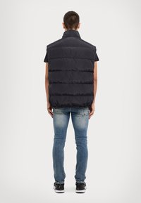 Gilet noir matelassé avec un col haut, design matelassé, sans manches, porté sur une chemise noire et un jean bleu, avec des baskets grises.