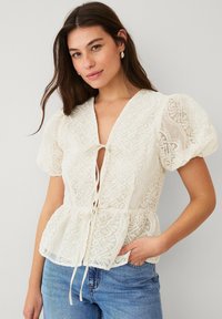 Hvid blondetop med puf ærmer, åben udskæring og snøreliv. Har broderede blomster mønstre og gennemsigtig stofdetaljering.