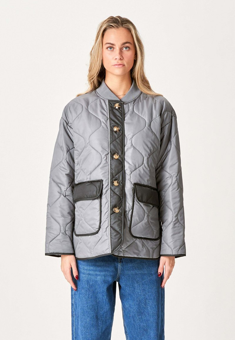 Grå quiltet jakke med rund krave, knaplukning foran og to store frontlommer med sort kant. Har guld knapper til lukning.