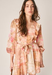 Junge Frau mit langen braunen Haaren trägt ein geblümtes Patchwork-Kleid mit Gürtel, Rüschendetails und langen Ärmeln, stehend vor einem schlichten Hintergrund.