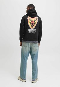 Schwarzer Hoodie mit einem bunten Grafikdesign eines Schädels und Flammen auf dem Rücken. Getragen mit hellblauen Jeans und beigen Schuhen.