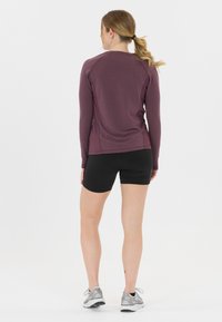 Sovitettu pitkähihaine violetti paita sileällä pinnalla, yhdistetty mustiin shortseihin ja hopeisiin urheilukenkään, nähtynä takaa.