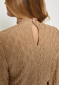 Beige, tekstureret bluse med diamantmønster. Har høj halsudskæring, nøglerum i ryggen og lukning med løkke. Blødt stof med synlige syninger.