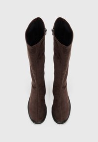 Une paire de bottes en daim brun foncé, hautes, avec des semelles noires, des tiges légèrement affaissées et des fermetures éclaires sur le côté, se tenant droit sur un fond blanc.