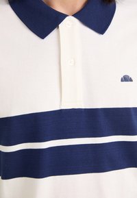 Polo blanc avec col bleu marine, deux bandes horizontales bleu marine séparées par une fine bande blanche, et petit logo bleu marine sur la poitrine.