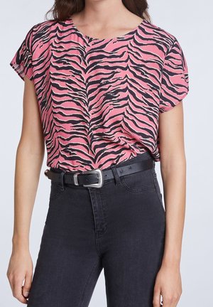 Femme portant un haut à manches courtes à imprimé zèbre rose et noir, rentré dans un jean noir taille haute avec une ceinture noire.