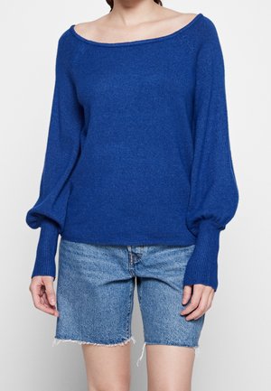 Vrouw draagt een blauw off-shoulder trui met ballonmouwen en lichtblauwe denim shorts met versnelde zoom.