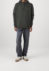 Uomo che indossa un felpa verde scuro con logo, jeans grigi larghi e scarpe da ginnastica bianche, in piedi su uno sfondo neutro.