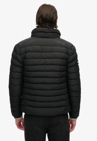 Sort pufferjakke med horisontal quiltet design, høj krave og ribbede manchetter. Har logo patch på overarmen. Materialet ser blødt og isolerende ud.