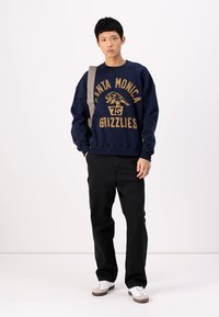Mornarska dukserica s natpisom "Santa Monica Grizzlies" u zlatnoj boji, uparena s crnim cargo hlačama i bijelim teniskama s bež detaljima.