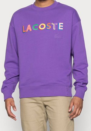Mand iført en lilla Lacoste sweatshirt med flerfarvede bogstaver på brystet og beige bukser mod en ensfarvet baggrund.