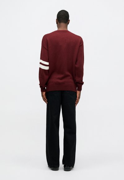 Bordå sant strikket genser med hvite striper på ermene, rund hals og løs passform. Kombinert med svarte vide bukser og svarte sko.