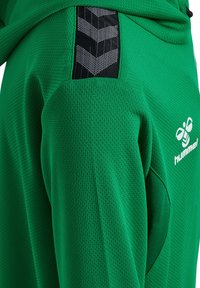 Grøn sportstrøje med tekstureret stof, der har sorte chevron-striber og en hvid logo. Designet fokuserer på åndbarhed og komfort.