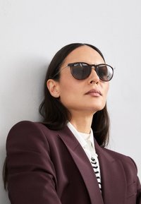 Ochelari de soare Ray-Ban cu lentile întunecate, ramă neagră și formă rotundă. Purtați de o persoană într-un sacou vișiniu peste un top alb cu dungi.