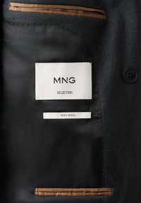 Interno di un blazer in lana nera con un'etichetta bianca che riporta "MNG SELECTION" e un'etichetta più piccola che indica "100% LANNA", accentuato da rifiniture marroni.