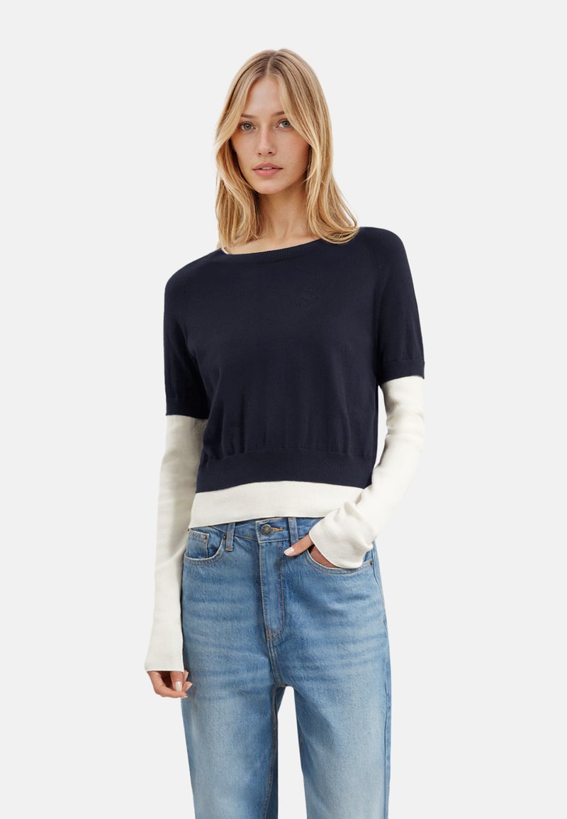 Maglione cropped blu navy con maniche bianche, realizzato in morbido tessuto a maglia, con scollatura rotonda e bordo a coste. Abbinato a jeans blu chiaro.