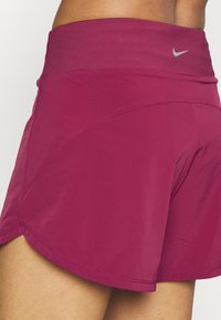 Bordeaux urheilushortsit, joissa on leveä vyötärönauha, joustava kangas ja kaareva helma. Sisältää pienen hopeisen Nike-logon vyötärön läheisyydessä.