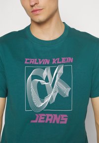 Turkusowa bawełniana koszulka z graficznym wzorem w białe i fioletowe abstrakcyjne linie, z napisem "CALVIN KLEIN JEANS" na dole.