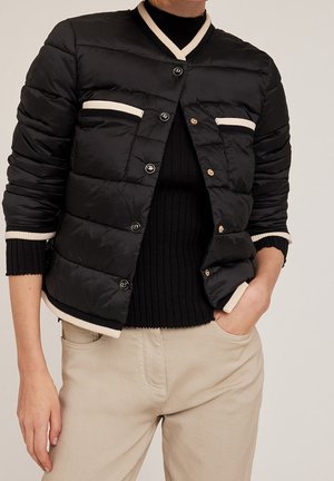 Veste matelassée noire avec bordure blanche portée sur un pull noir côtelé, associée à un pantalon beige, main du mannequin dans la poche et fond neutre.