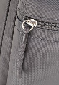 Borsa in tessuto grigia con superficie testurizzata, dotata di una tasca con zip e un'etichetta in tessuto con un accento di anello metallico.
