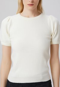 Pull en tricot blanc à manches courtes et col rond. Doté de manches bouffantes et d'une bordure côtelée au niveau du col et de l'ourlet. Texture douce.