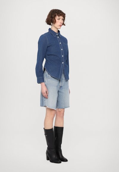 Chemise en denim à boutons avec des manches longues et une poche à l'avant, associée à un short en denim bleu clair et des bottes noires à talons hauts jusqu'aux genoux.