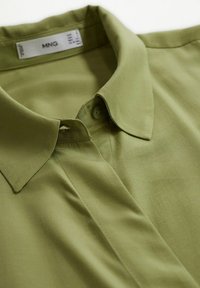 Chemise vert olive à col avec patte de boutonnage cachée, texture de tissu lisse, et étiquette de marque à l'intérieur du col.