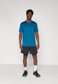 Blå trænings t-shirt med korte ærmer, parret med sorte shorts. Modellen bærer sorte sneakers og hvide sokker, stående på en neutral baggrund.